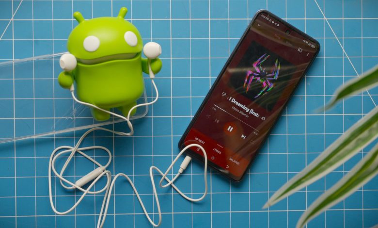 Tecno Pova 5 Pro playign YouTube Music with included wired earbuds and Android figure